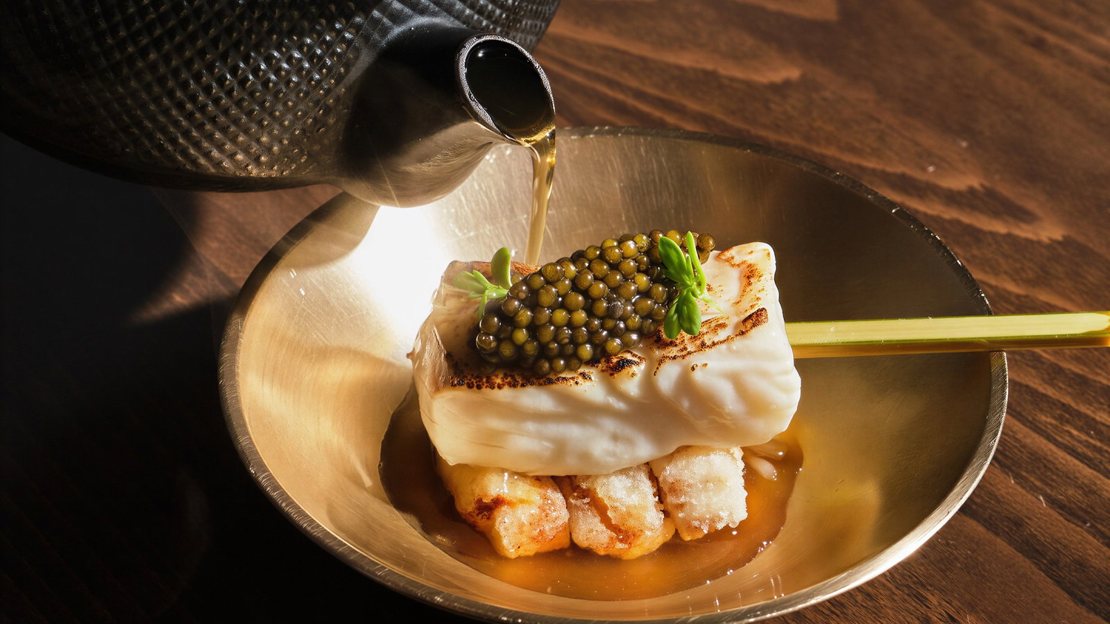 A black teapot pours broth over a dish with caviar on whipped cream, set on a wooden table. The mood is elegant and refined.