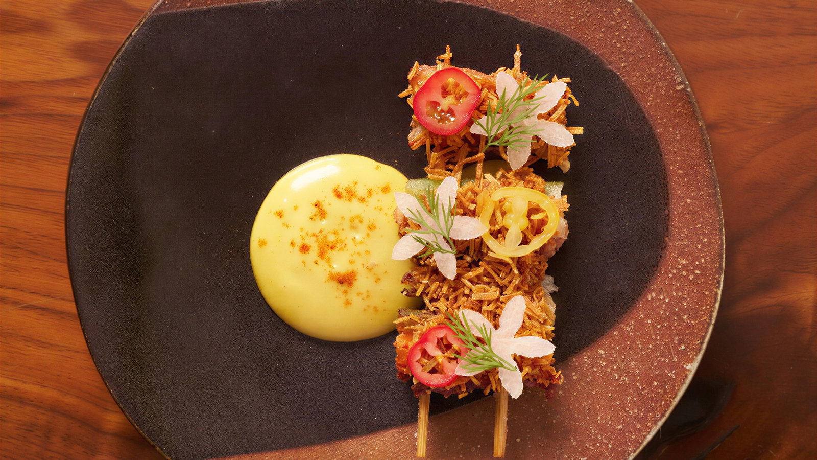 Elegant dish on black plate with skewers topped with sliced chili, white flowers, herbs, beside a creamy yellow sauce, on wood table.