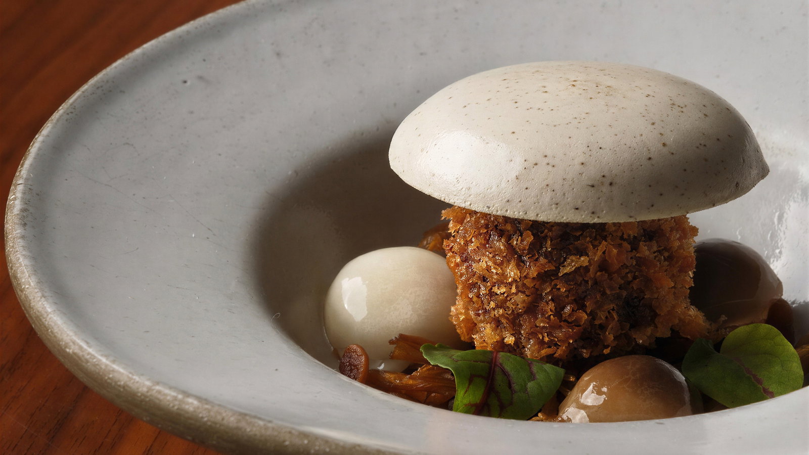 Gourmet dish with a crispy ball, white foam cap, and leaves on a gray plate. Wood table background. Elegant plating in warm tones.
