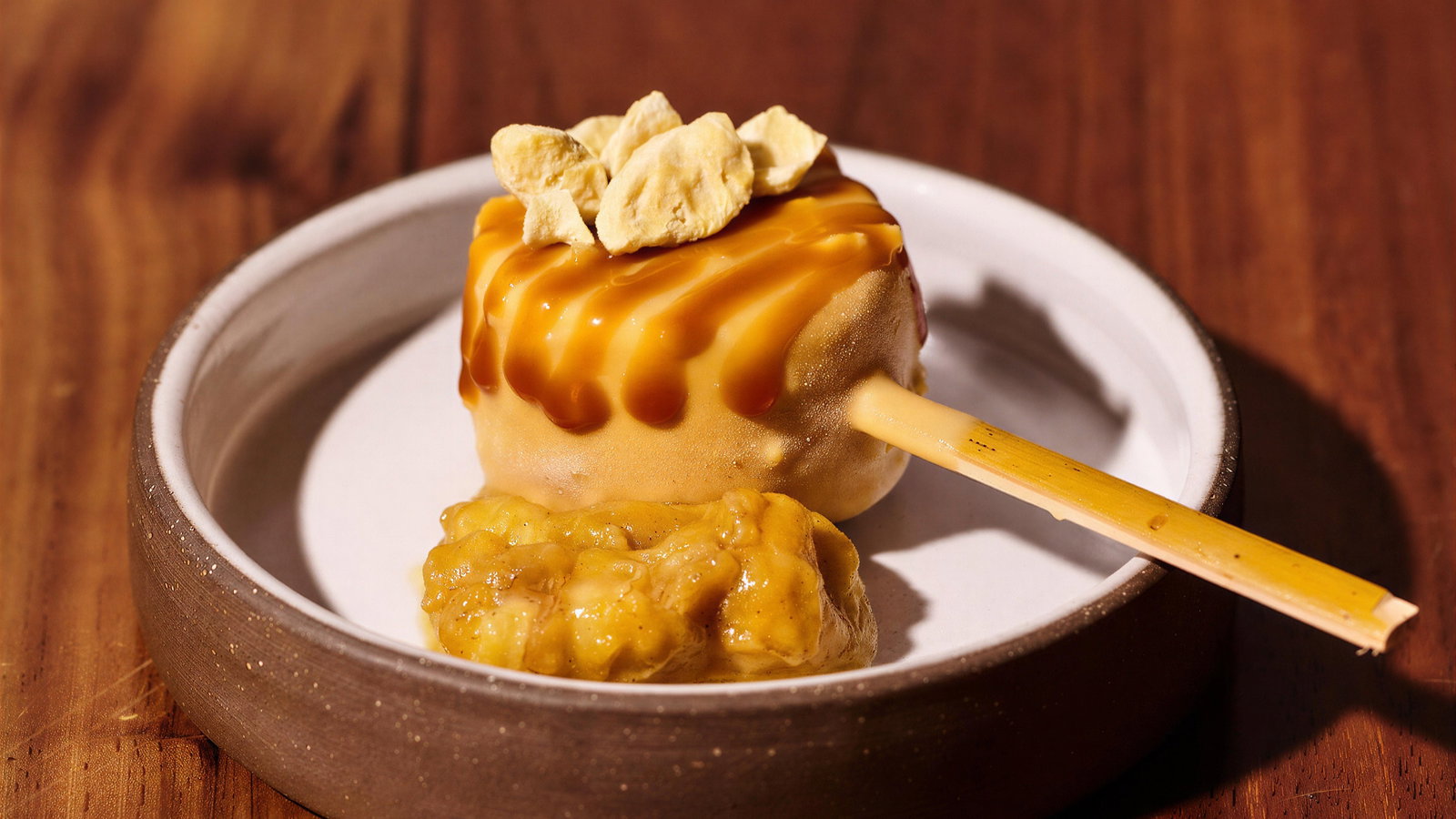 Ice cream on a stick, drizzled with caramel and topped with nuts, sits in a white bowl on a wooden table, evoking a delicious mood.