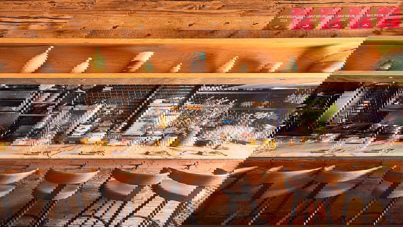 Rustic restaurant interior with a wooden bar, stools, and black-tile kitchen. Shelves hold vases. Four Michelin plaques on the wall.