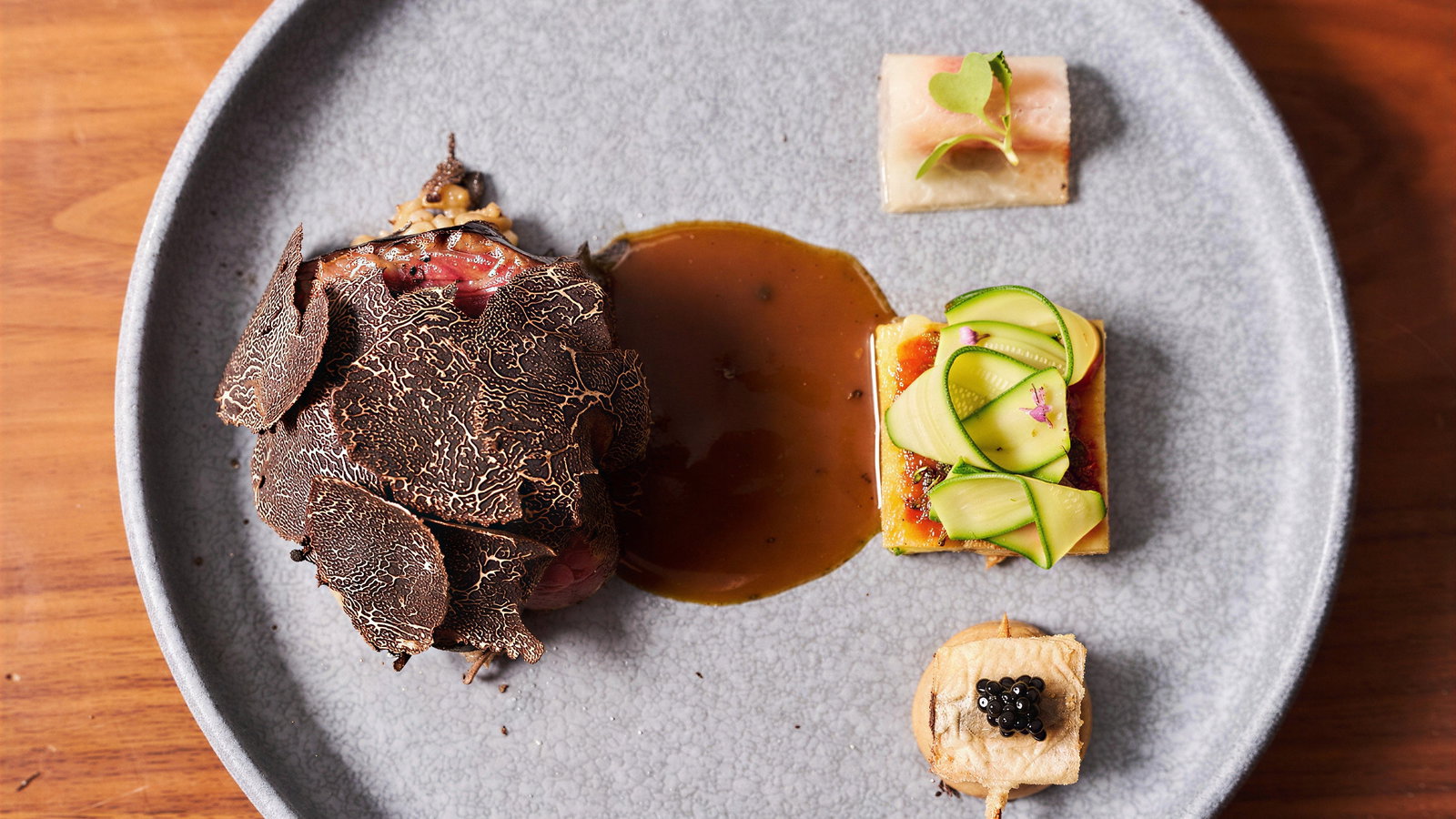 Gourmet dish on a gray plate: truffles, sauce, zucchini slices, and caviar. Set on a wooden table, showcasing elegant plating.
