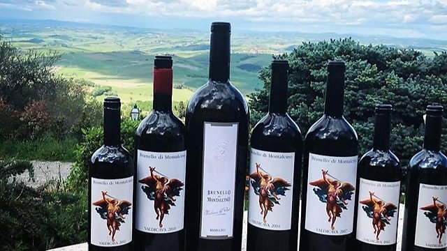 Seven Valdicava wine bottles labeled "Brunello di Montalcino" on a stone table, with a scenic green landscape and cloudy blue sky in the background.