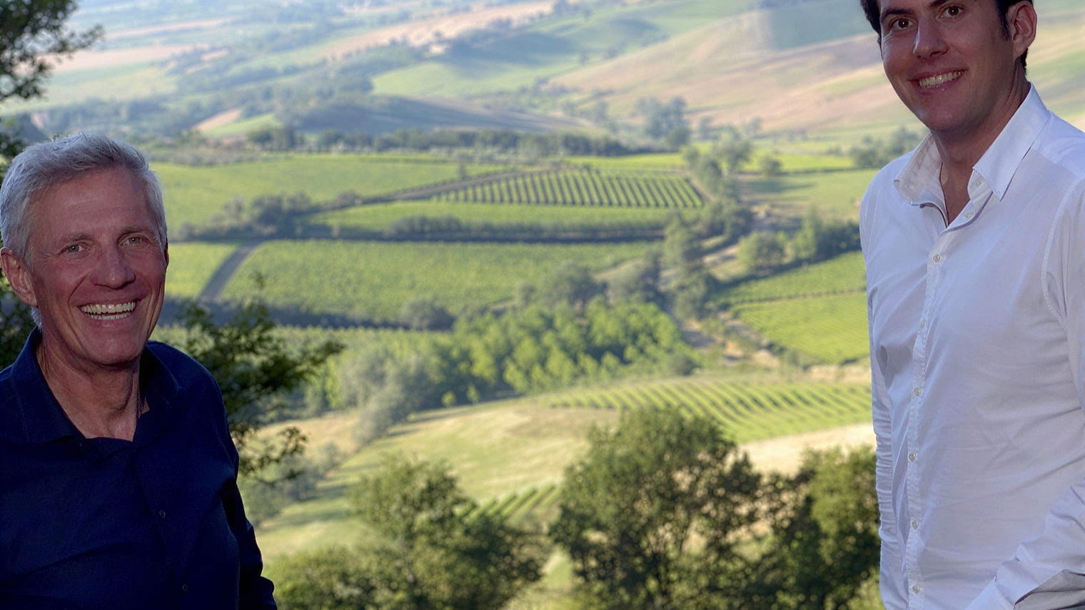 Valdicava Winemaker Vincenzo Abbruzzese and his son Pierfilippo smiling in front of a scenic landscape with rolling green fields and trees. One wears a blue shirt, the other a white shirt.