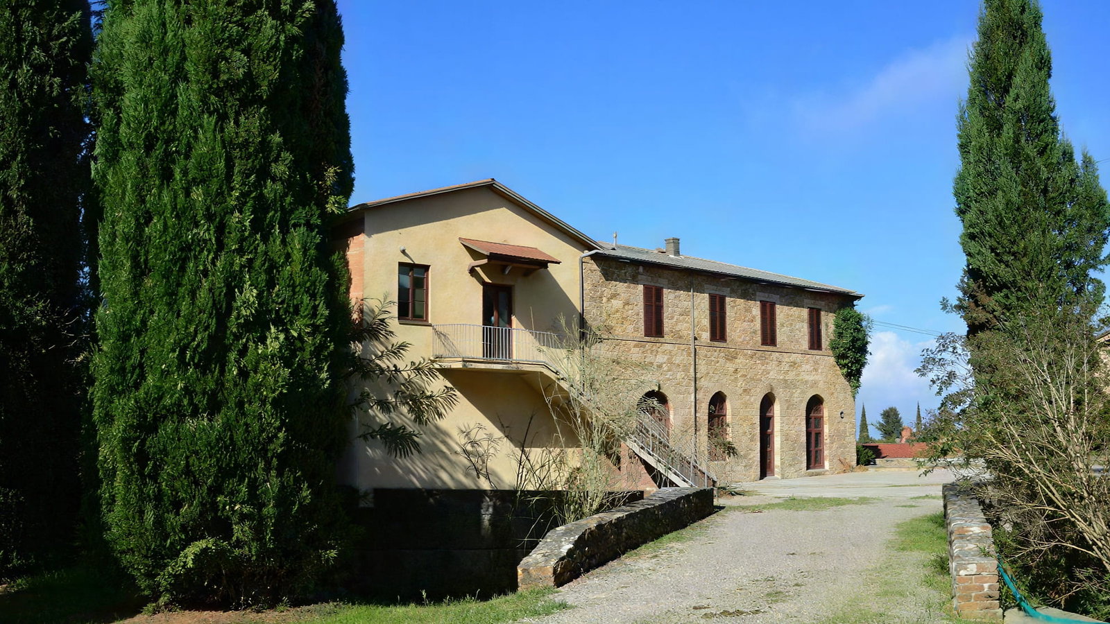 Stone farmhouse estate of Valdicava with red shutters, surrounded by tall cypress trees under a clear blue sky. Gravel path leads to the entrance. Tranquil setting.