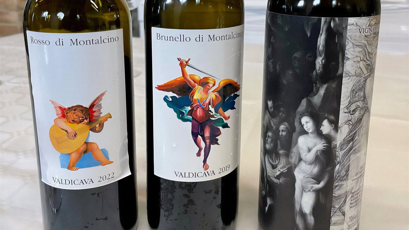 Three Valdicava wine bottles labeled Rosso di Montalcino 2022, Brunello di Montalcino 2019, and another with artistic imagery, on a table with a stone wall.
