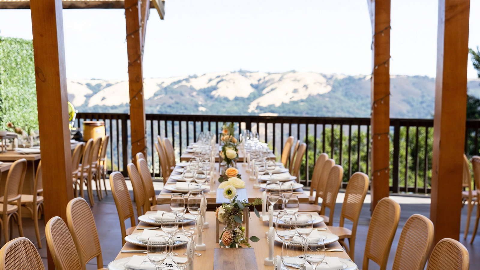 Elegant outdoor dining setup with neatly arranged tables. Wooden pergola, mountain views, orange roses, and wine glasses create a serene atmosphere.