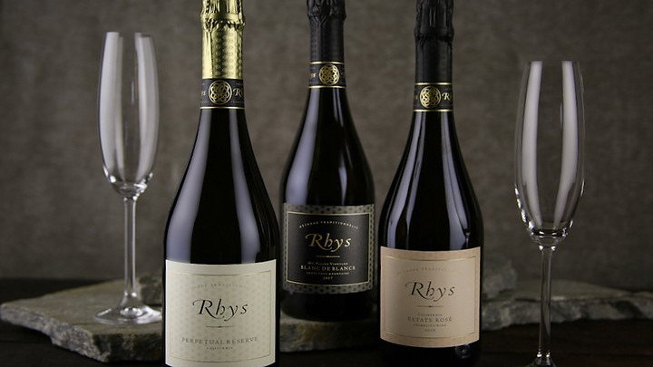 Three Rhys wine bottles centered between two empty champagne flutes on a stone surface. Labels read "Perpetual Reserve," "Blanc de Blancs," "Estate Rosé."