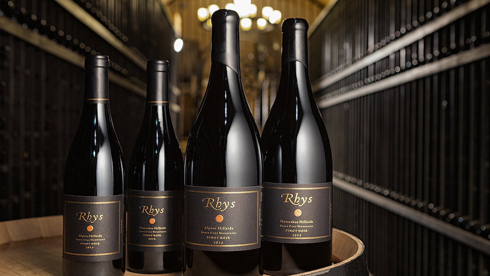 Four Rhys wine bottles sit on a barrel in a dimly lit cellar. The labels read Pinot Noir, vintage 2013. Dark, elegant setting.