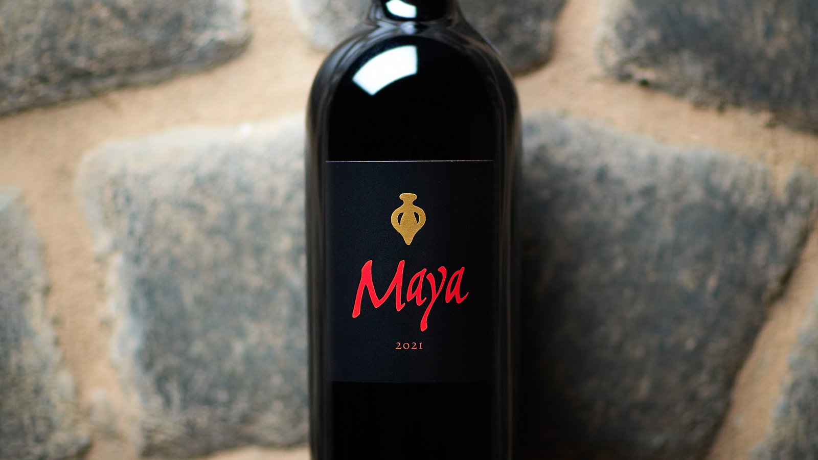 A wine bottle labeled "Maya 2021" with red text on black, set against a cobblestone background. The bottle has a gold emblem.