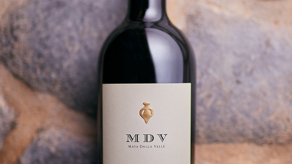 A wine bottle labeled MDV 2018 stands against a textured stone backdrop. The label is cream-colored, creating an elegant contrast.