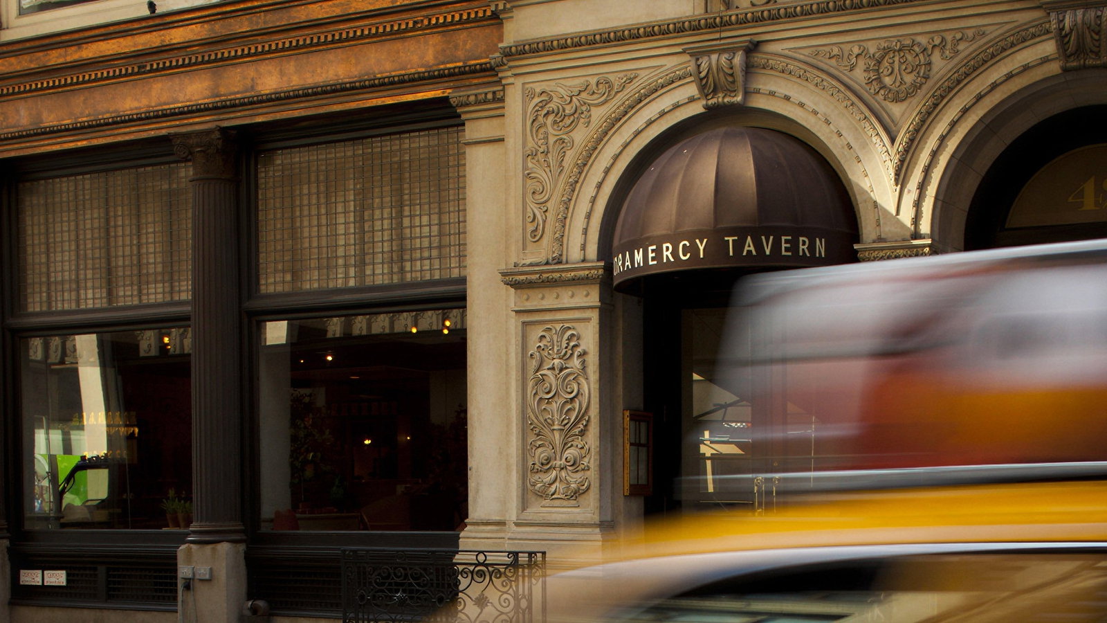 Elegant city building facade with Gramercy Tavern entrance, blurred yellow taxi passing by, creating a dynamic, urban atmosphere.