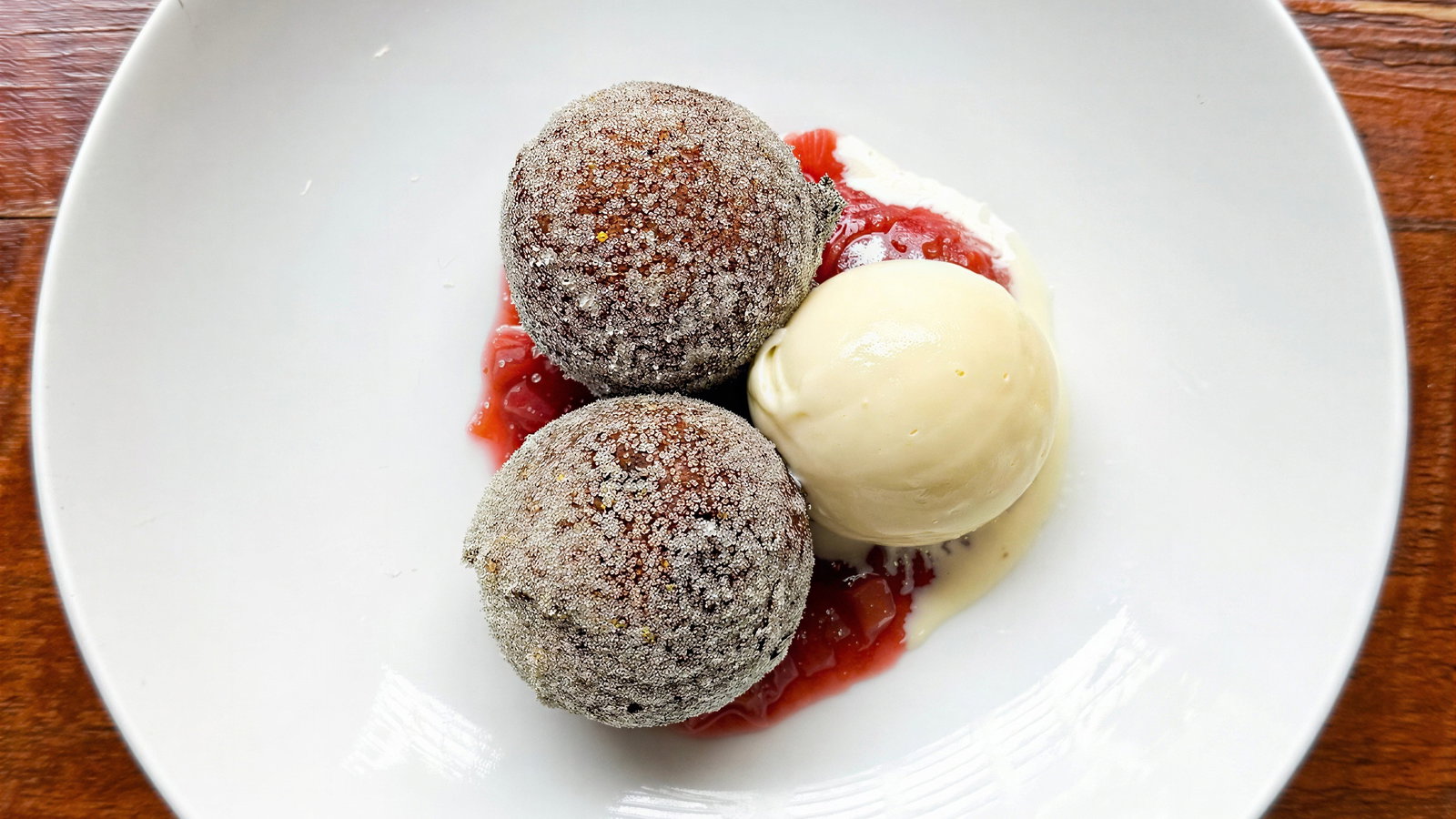 Two sugared doughnuts with a scoop of vanilla ice cream and red fruit sauce on a white plate. Wooden table visible beneath.