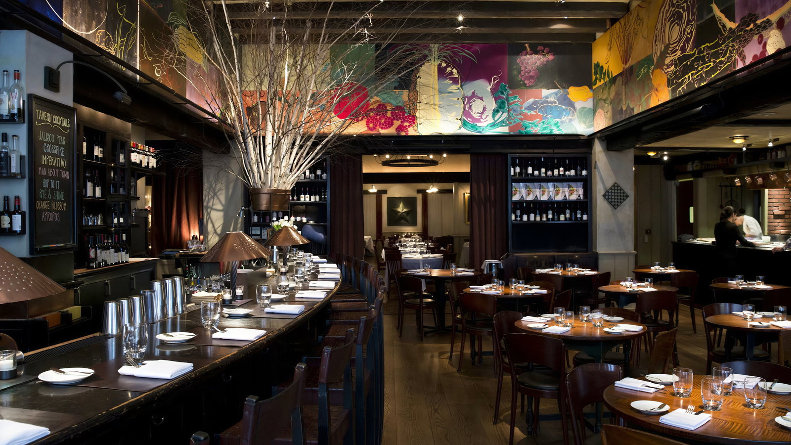 Dimly lit restaurant with wooden tables, colorful mural, and large bar. Bottles line shelves; two chefs work in the kitchen. Cozy ambiance.