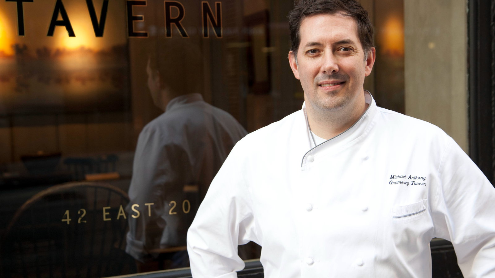 Chef in white uniform smiles outside a restaurant with "TAVERN" and "42 EAST 20" on window. Reflective glass shows dim interior.
