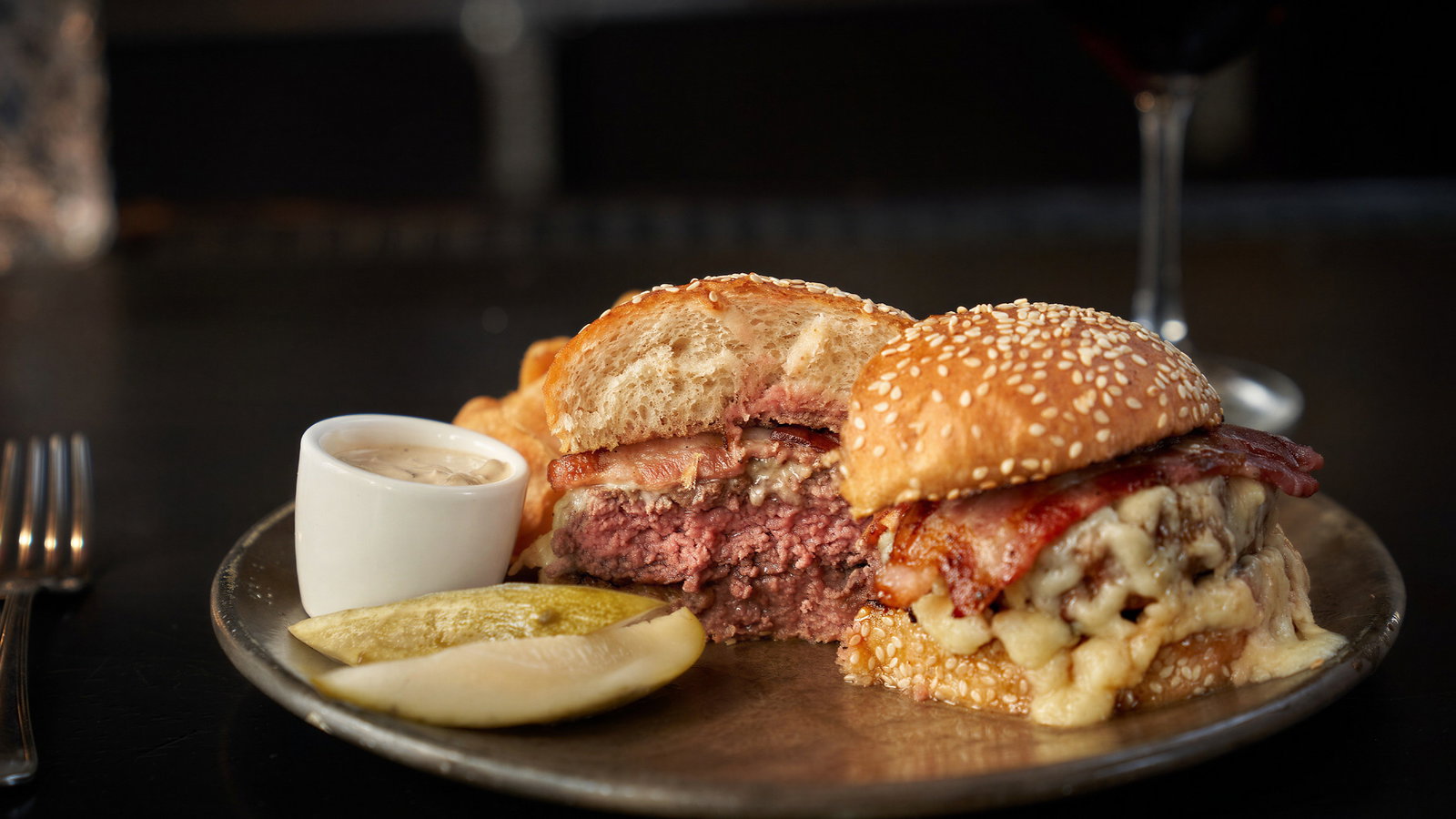 Cheeseburger with bacon on a sesame bun, cut in half on a plate with pickles and sauce. Fork and red wine glass in background.