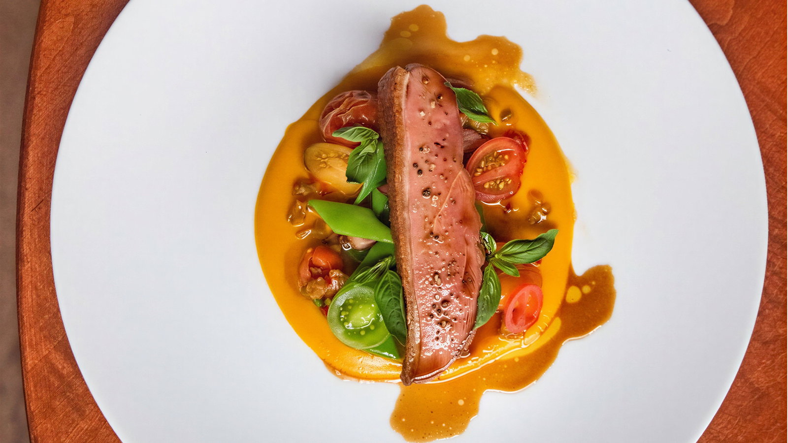 Gourmet dish on a white plate: seared meat atop vibrant orange sauce, garnished with cherry tomatoes and herbs, on a wooden table.