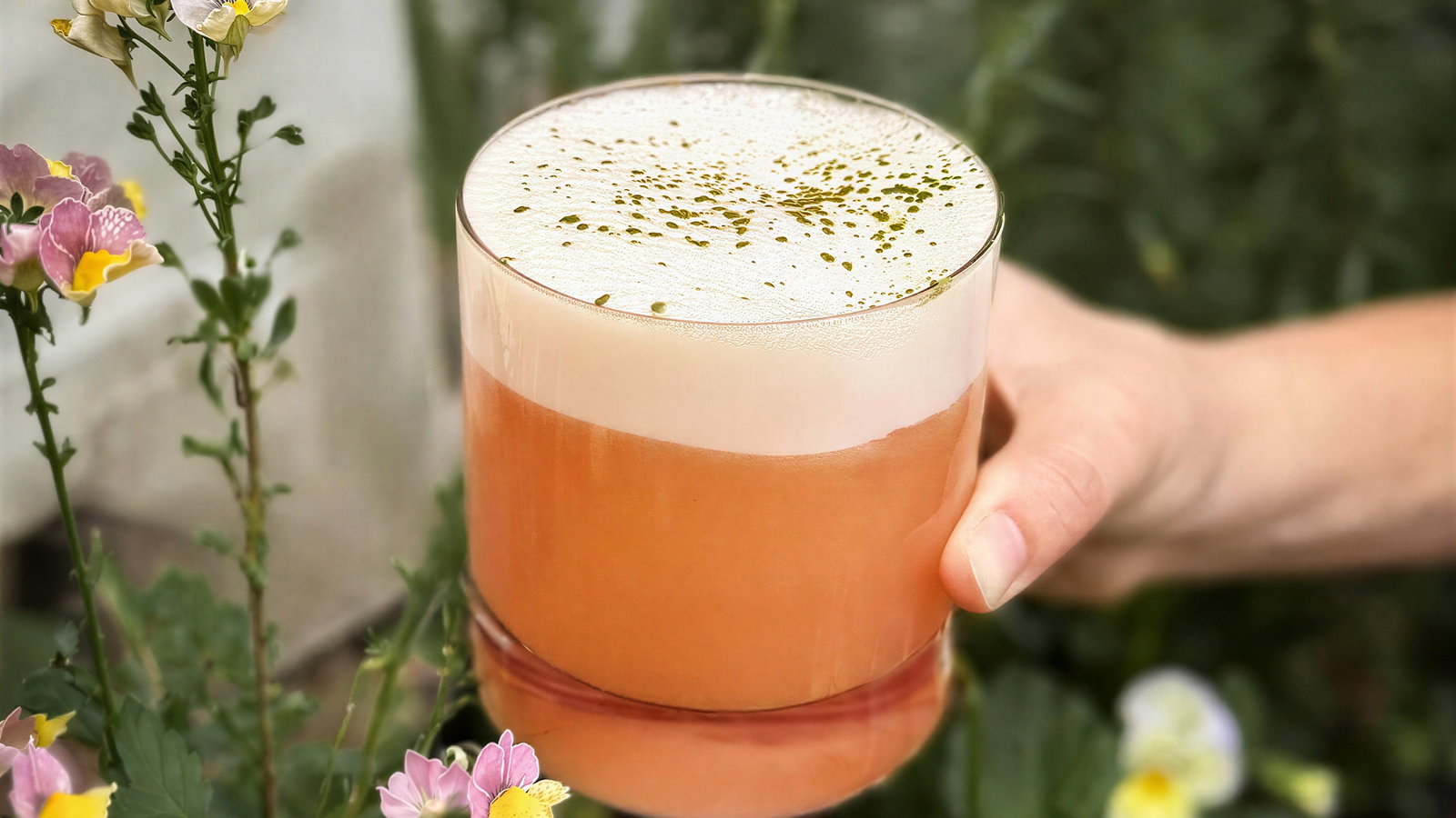 A hand holds an orange drink with frothy top, sprinkled with green bits, next to pink and yellow flowers. Background is lush greenery.