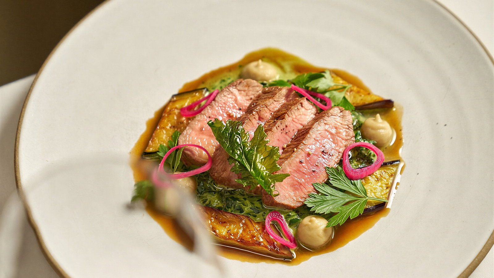 Gourmet dish of sliced pink lamb on a white plate with colorful garnishes, greens, and sauce. Glass of red wine in foreground. Elegant setting.