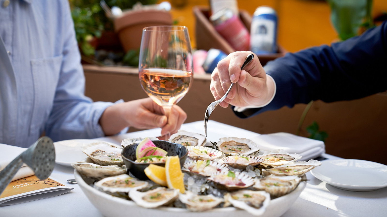 Two people enjoy a platter of oysters with lemon slices, one holding a fork and another a glass of rosé. Outdoor dining, relaxed vibe.