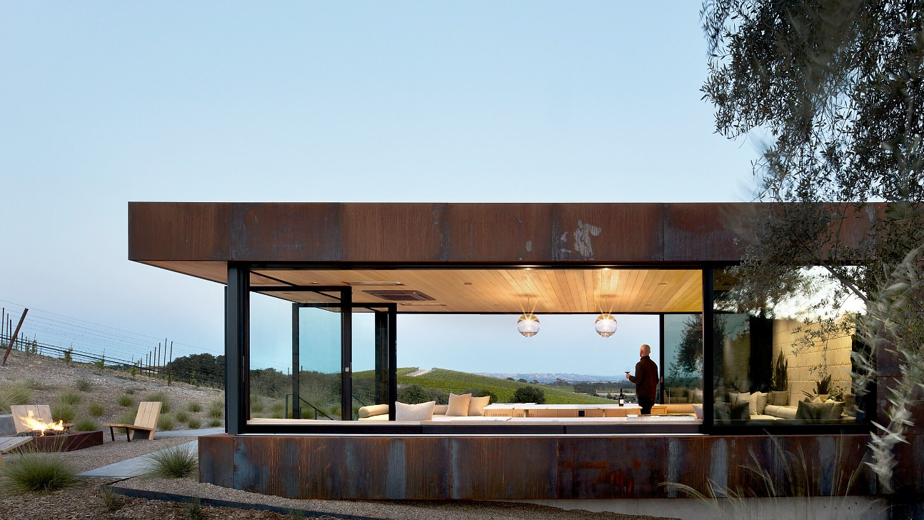 Modern house with large glass windows framing a scenic view. A person stands inside with wine. Rustic exterior, serene ambiance.