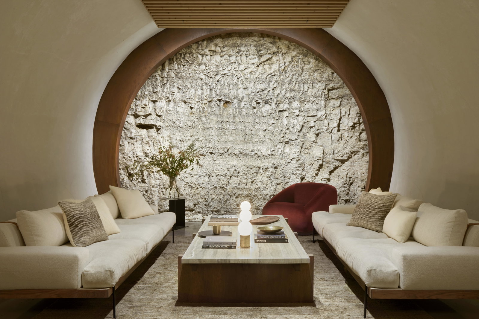 Modern living room with beige sofas, red armchair, stone wall, and wooden accents. Central marble table holds books and decor. Cozy ambiance.