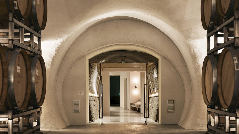 Stone wine cellar with rows of wooden barrels on each side, lit warmly. An entrance to a modern room is visible in the background.