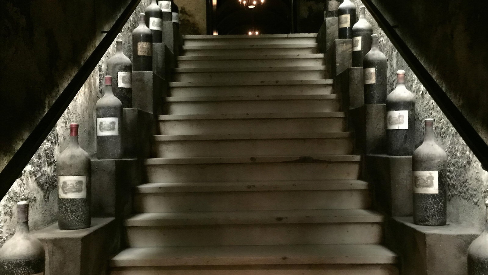 Dimly lit stone staircase lined with large, dusty wine bottles on either side. Dark, rustic ambiance with chandeliers visible in the distance.