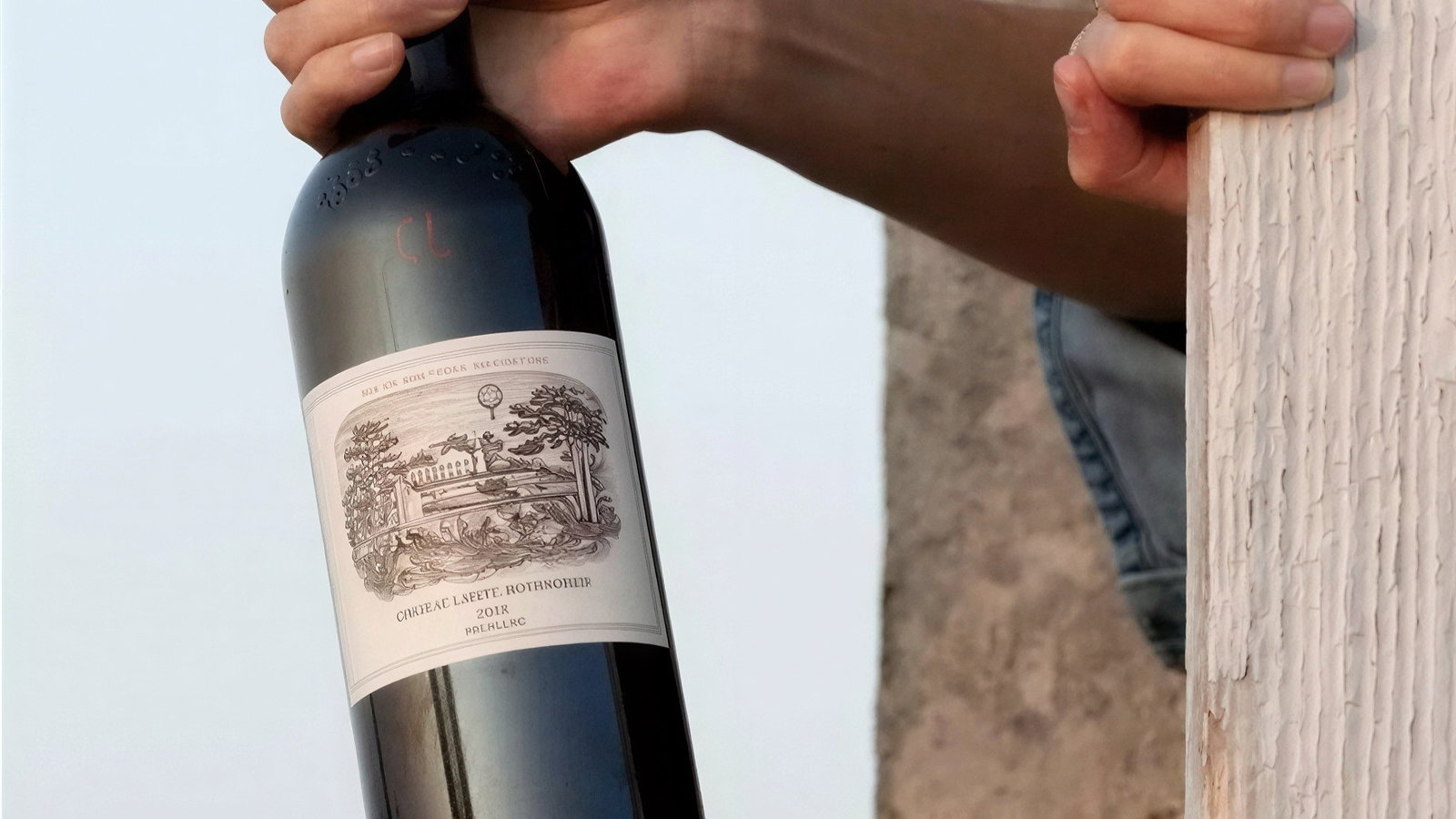 A hand holds a wine bottle labeled "Château Lafite Rothschild 2017." Background shows stone and wood, set against a clear sky. Mood is serene.