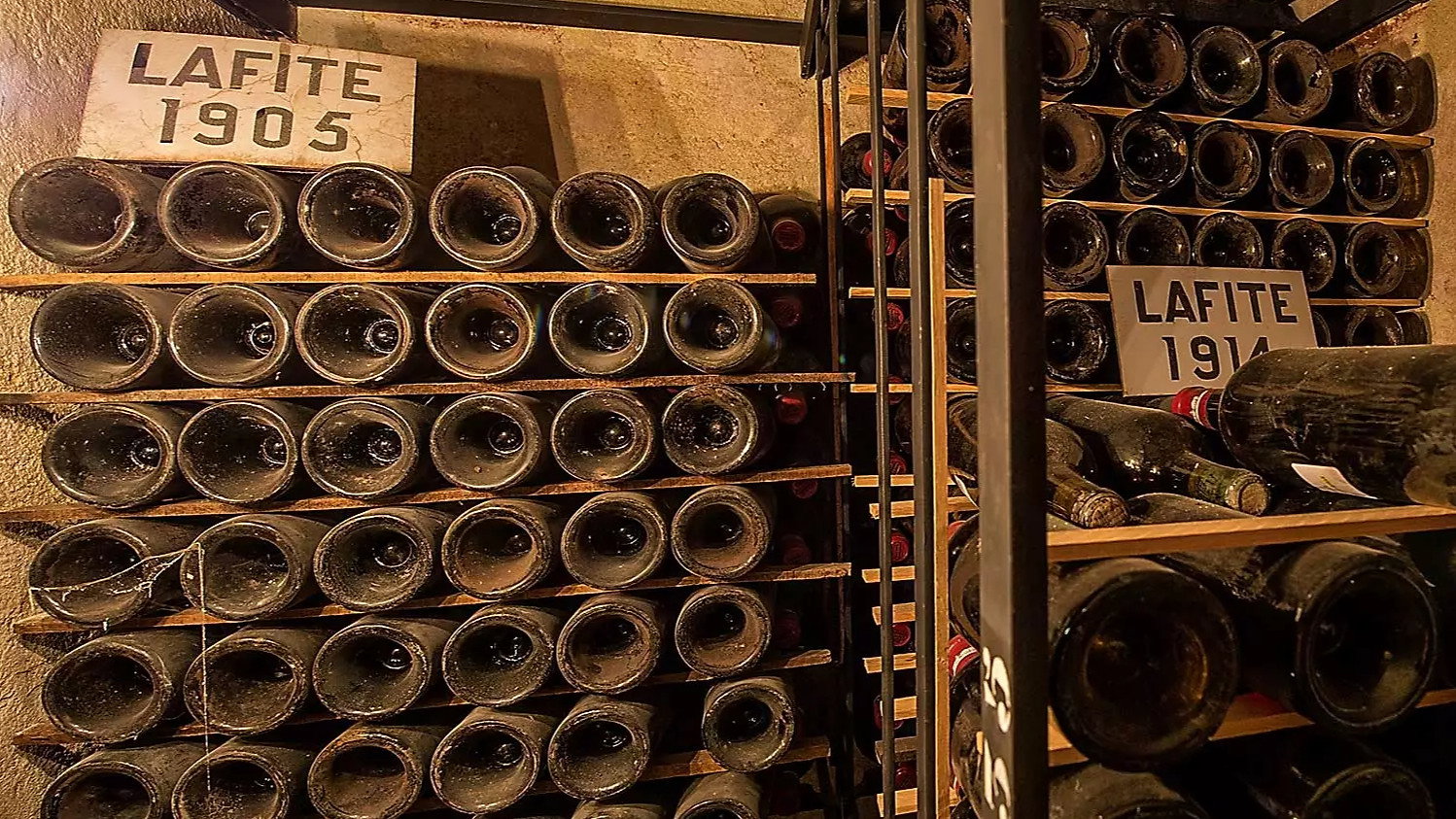 Wine bottles on wooden racks in a dim cellar. Labels read Lafite 1905 and 1914. Vintage ambiance with aged, dusty bottles.