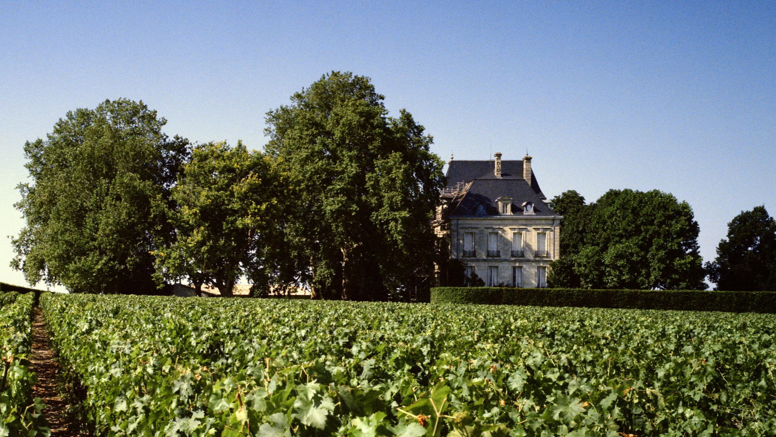 A grand French chateau with stone facade, slate roof, and multiple windows. A manicured garden and steps lead to the entrance.