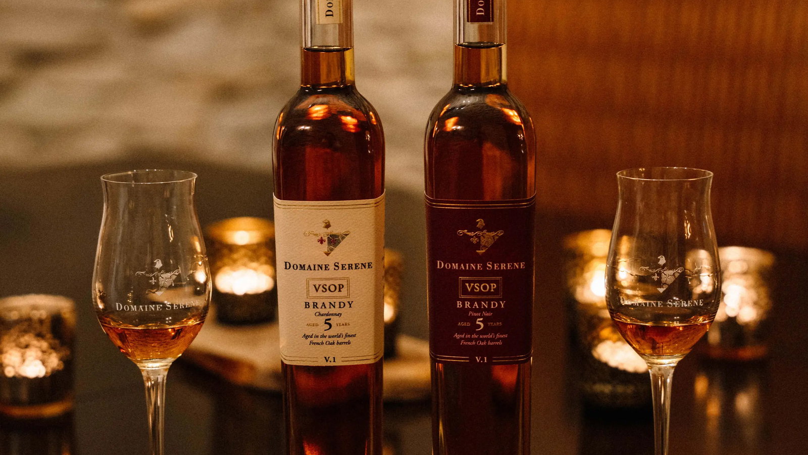 Two Domaine Serene VSOP brandy bottles and glasses on a dark table. Warm, ambient lighting from candles creates a cozy atmosphere.
