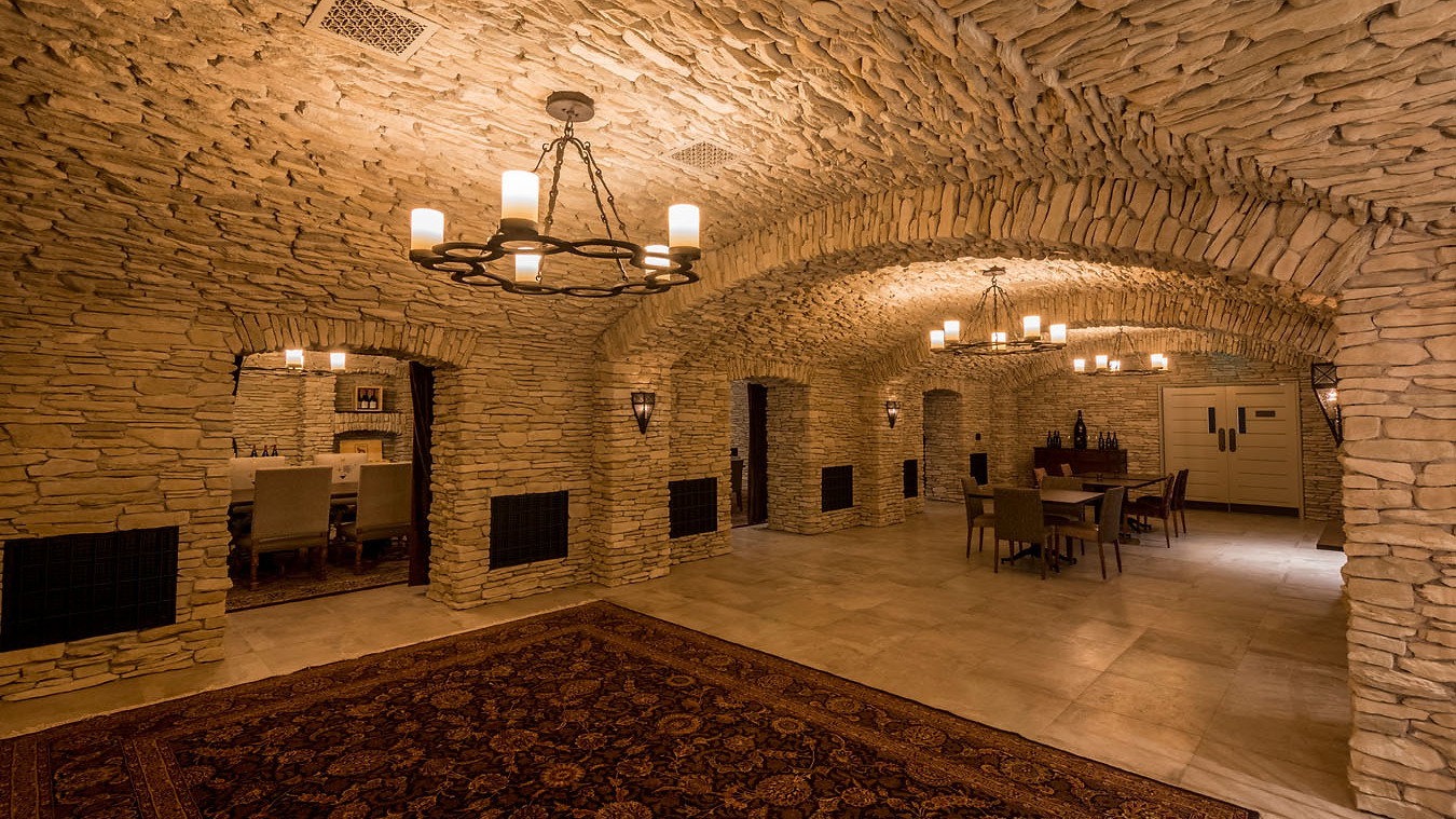 Stone-walled wine cellar with candle chandeliers, a richly patterned rug, and wooden tables; dimly lit, cozy ambiance.