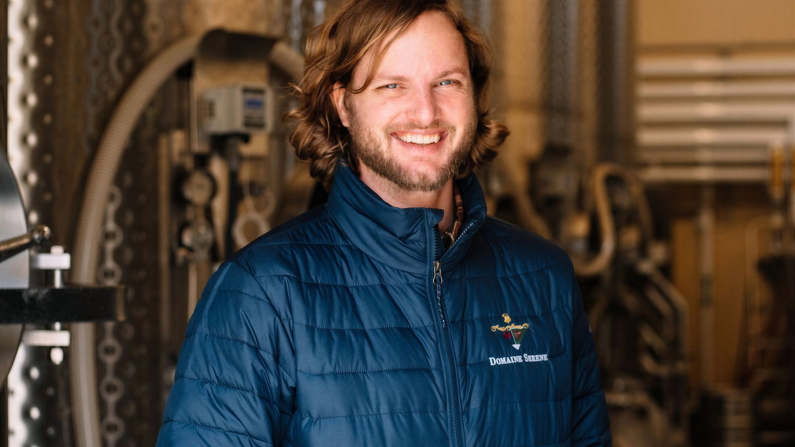 Smiling man in blue jacket stands in an industrial setting with large tanks. Jacket reads "Domaine Serene." Warm, inviting mood.