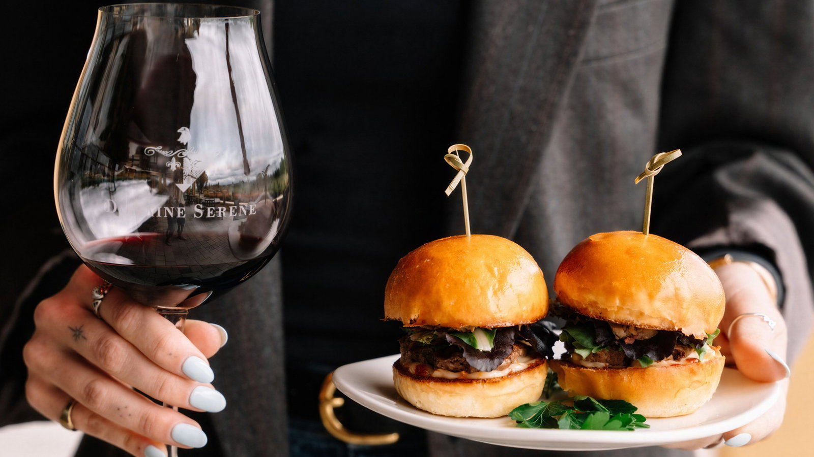 Person holding a wine glass with "Domaine Serene" text and a plate with two sliders. Dressed in a dark blazer, casual setting.
