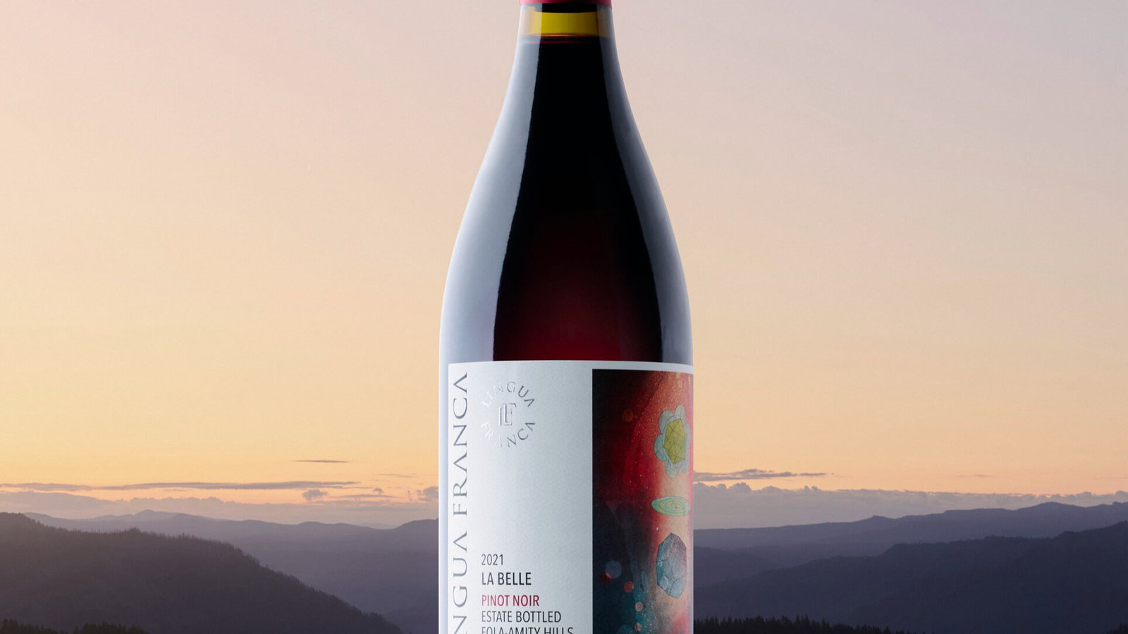 Red wine bottle labeled "Lingua Franca La Belle 2021" on reflective surface. Mountains and soft pastel sky create a serene background.