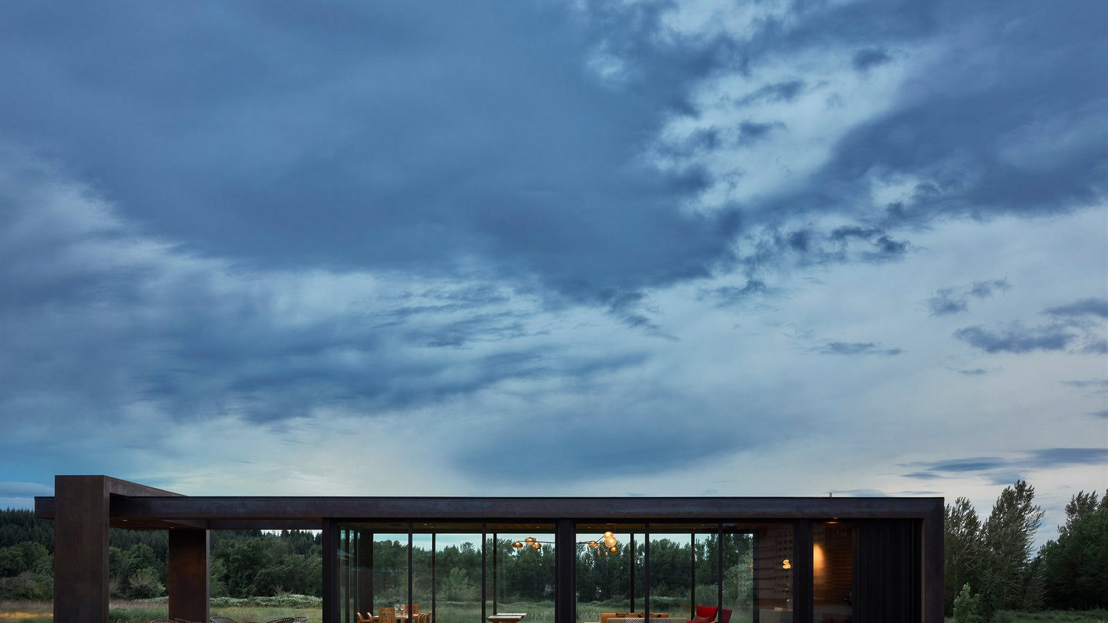 Modern glass house with warm interior lighting set in a lush green landscape under a dramatic cloudy sky. Cozy and serene atmosphere.