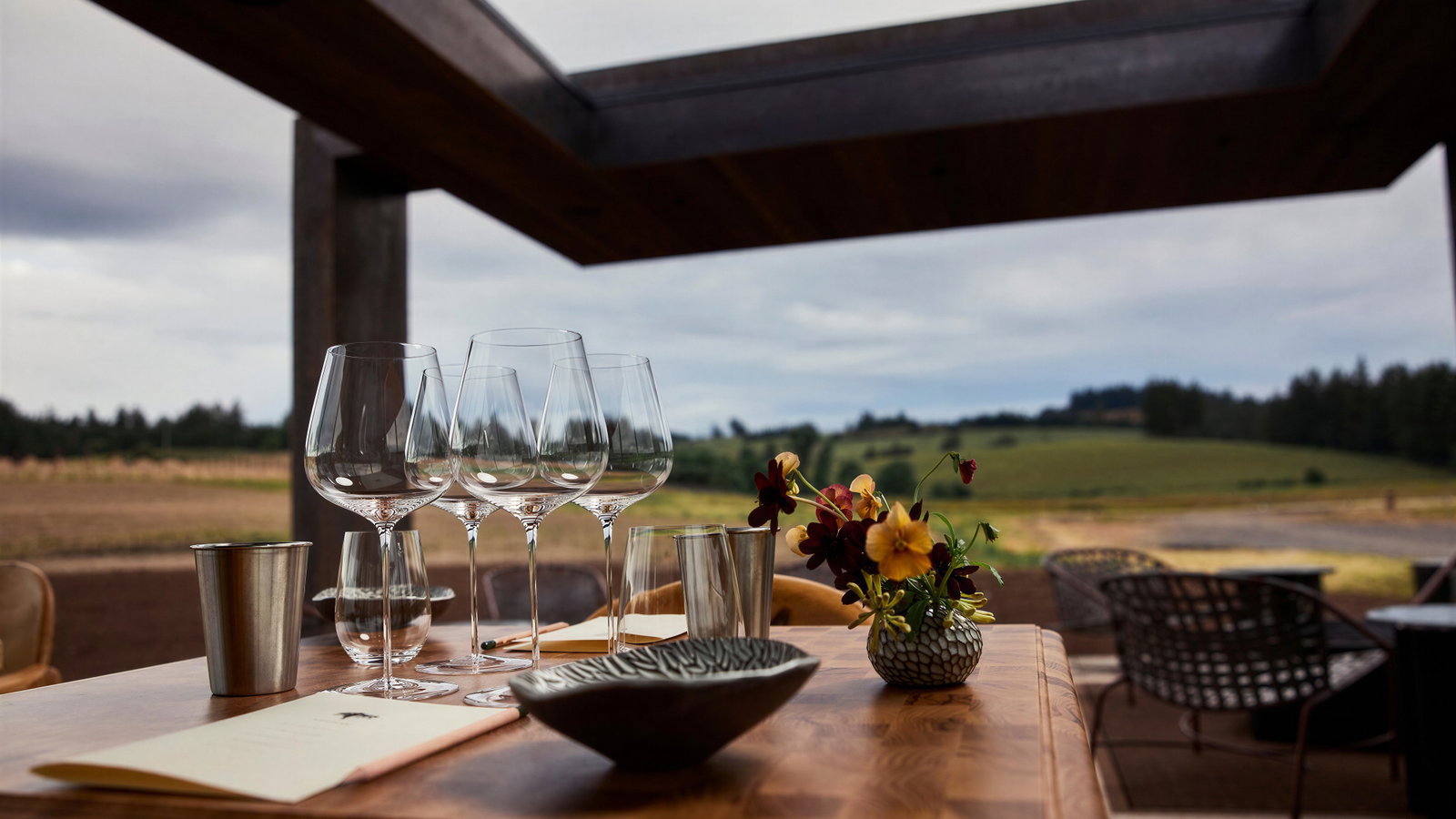 Table with empty wine glasses, a steel cup, and a flower vase on a patio overlooking green fields under a cloudy sky. Cozy, calm setting.
