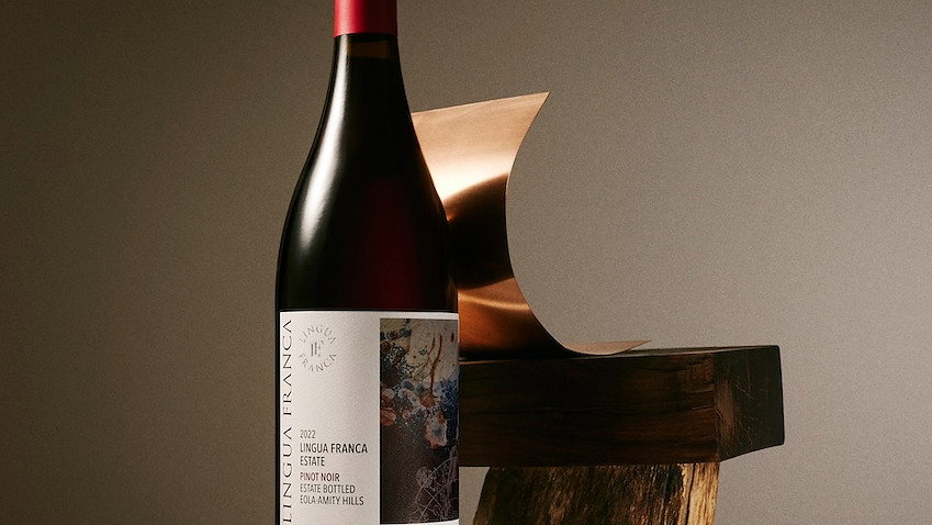 A bottle of red wine with a red cap titled 2022 Lingua Franca Pinot Noir sits on rustic wood against a gray background.