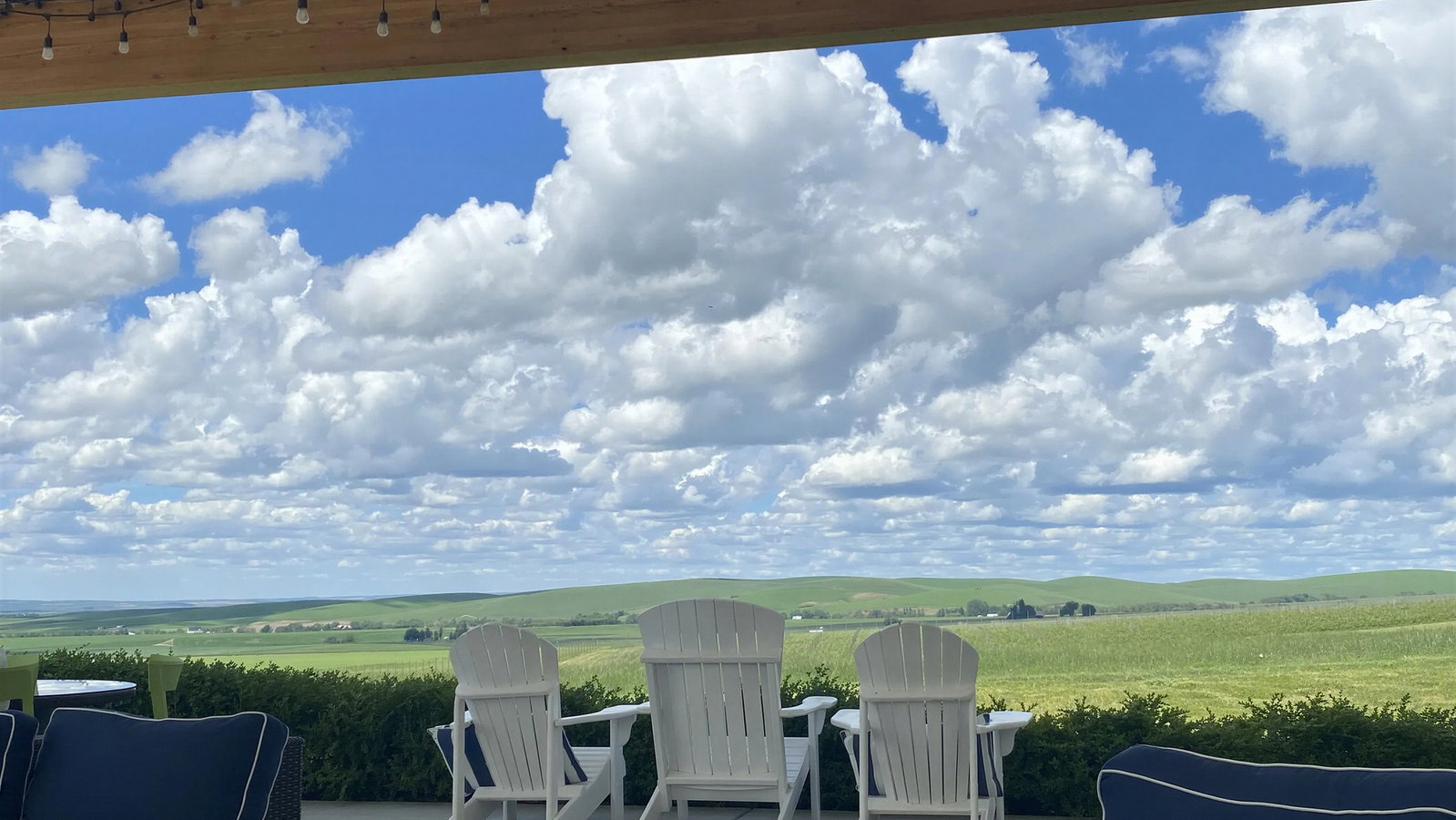 Patio with white chairs facing vast green fields under a blue sky with fluffy clouds. String lights hang above, creating a serene atmosphere.