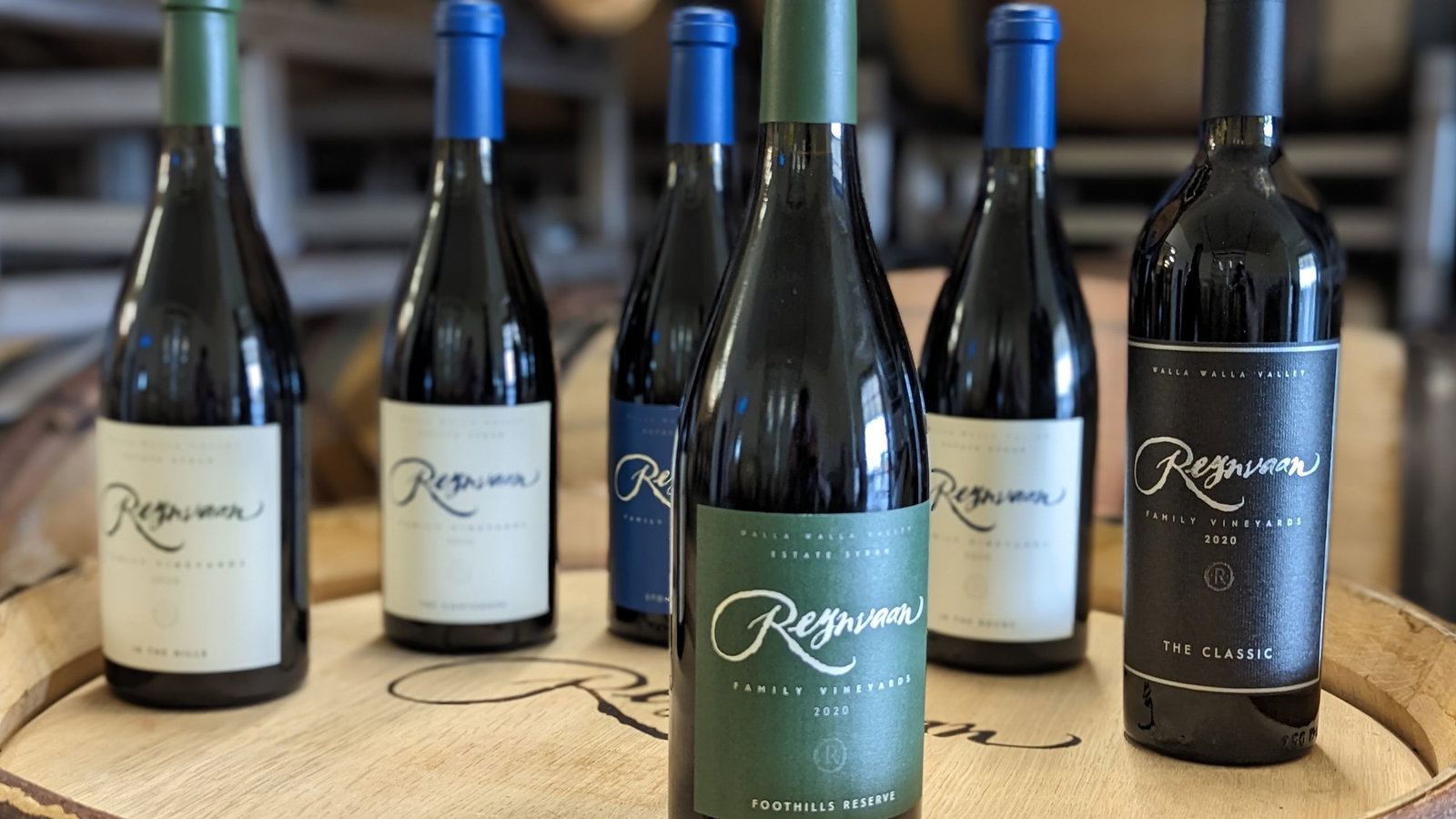 Five wine bottles with varied labels sit on a wooden barrel in a winery. Labels read "Rasa" and "Resqwaan Reserve." Background has wine barrels.