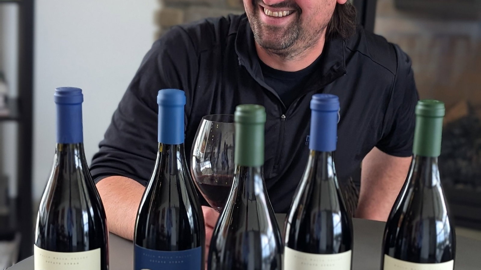 Smiling man in a dark beanie holds wine glass, surrounded by five wine bottles labeled "Rezman." Stone fireplace in background. Cozy setting.