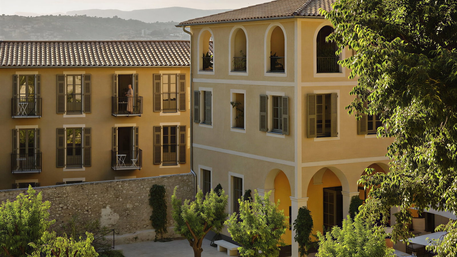 Exterior of Hôtel du Couvent yellow buildings with arched windows, balcony with a person, trees in a sunny courtyard. Hills in the background create a serene mood.