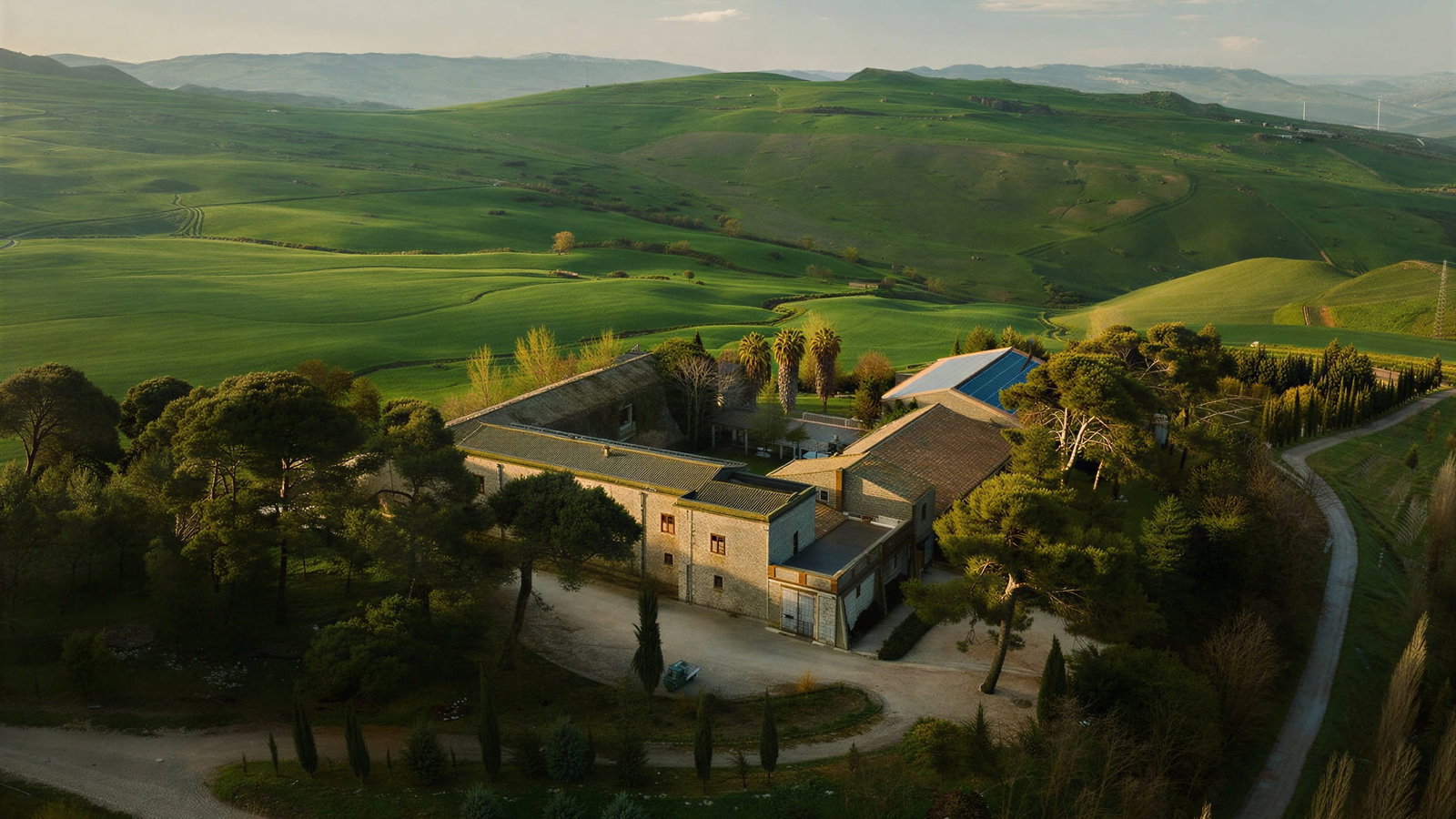 Aerial view of Masseria Susafa surrounded by lush green hills and trees under a partly cloudy sky. Winding dirt road visible. Calm mood.