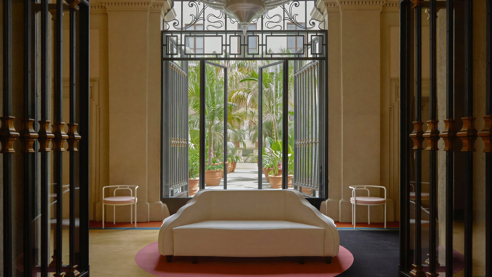 Palazzo Talìa entrance room with white sofa, colorful geometric rug, and large chandelier. Open glass doors reveal a lush garden outside.